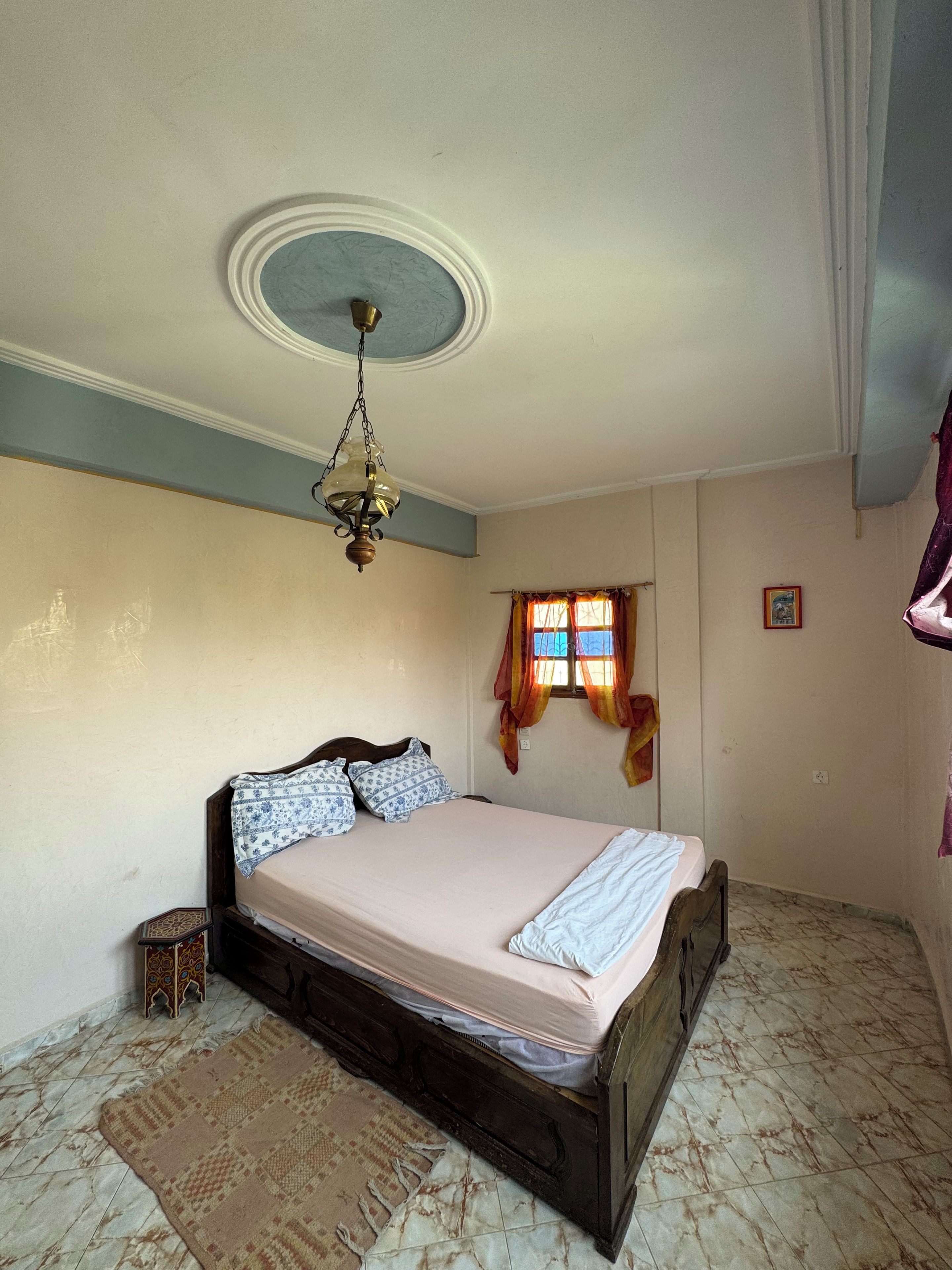Un dormitori amb un llit, una làmpada penjant, una finestra amb cortines i un objecte decoratiu petit al terra.