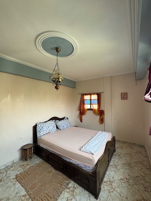 A bedroom with a bed, a hanging light fixture, a window with curtains, and a small decorative item on the floor. - Welcoming 2-bedroom apartment in vibrant Aourir perfect for relaxing stay (Agadir)