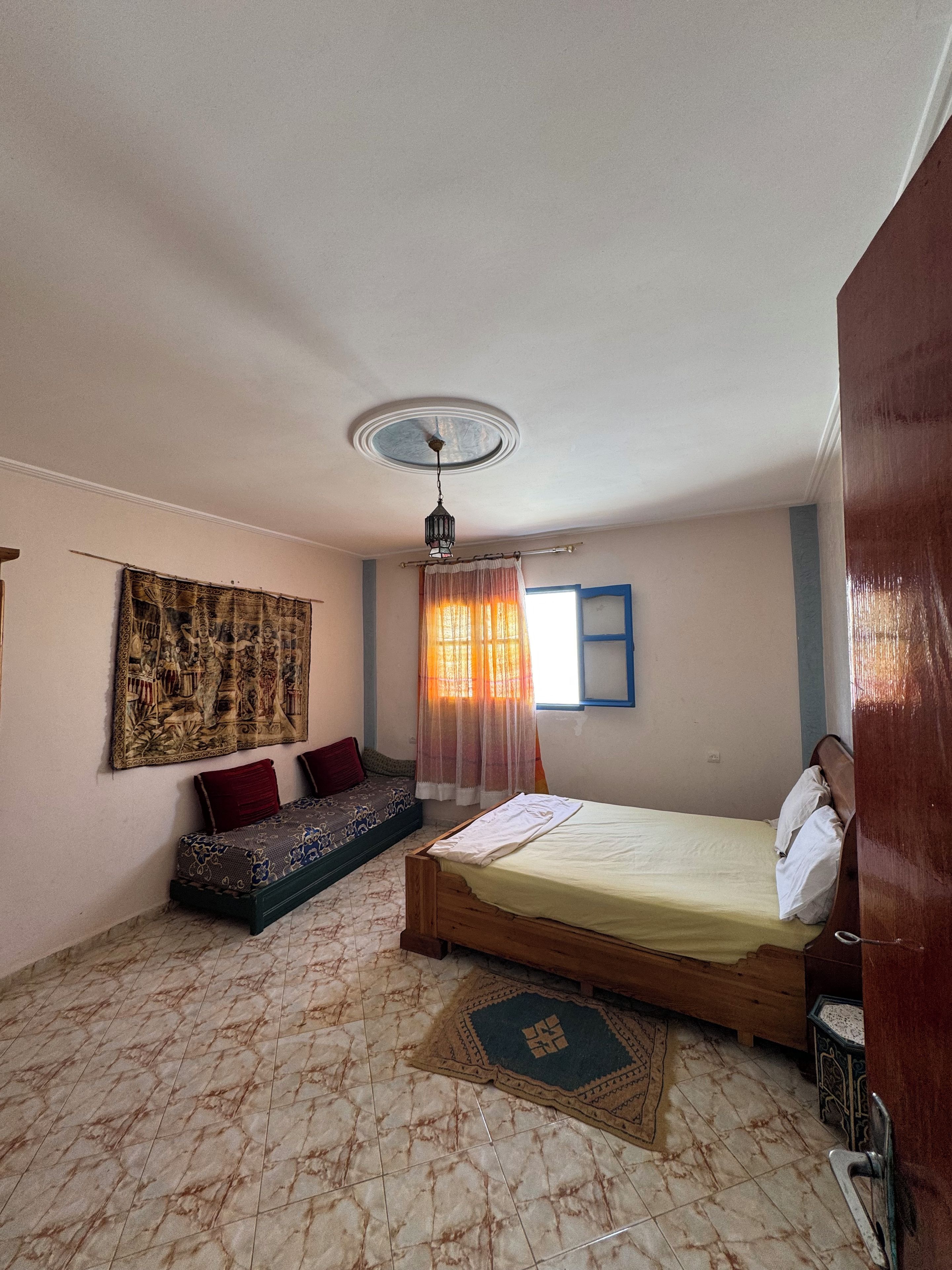 A bedroom with a bed, a bench, a ceiling fan, a window with curtains, and a tapestry on the wall.