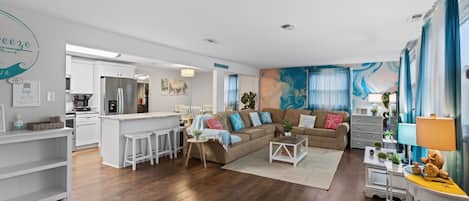 A modern living room with a sofa, coffee table, and a kitchen area with a refrigerator and bar stools.