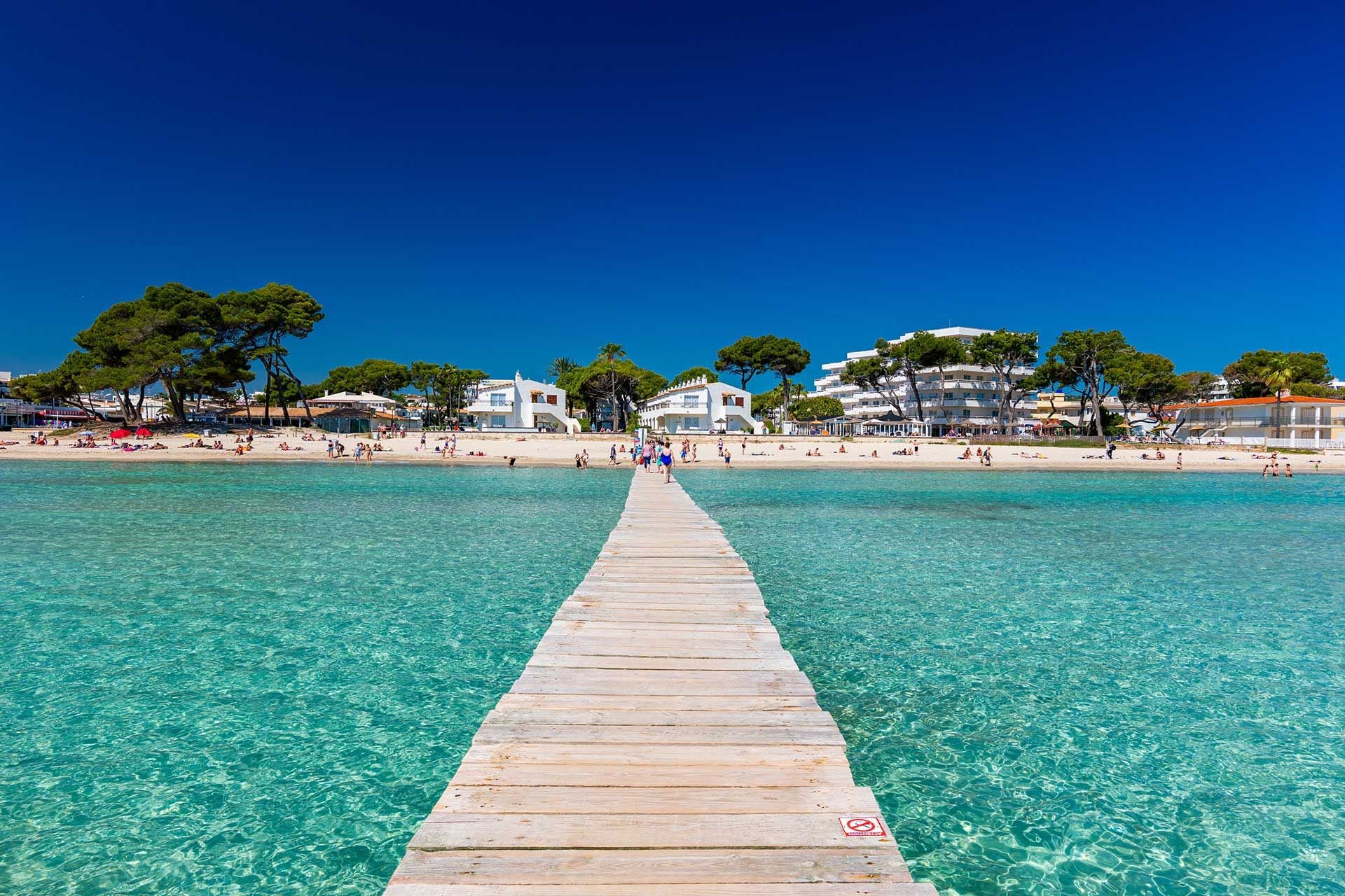 A wooden pier leading to a beach with clear turquoise water, surrounded by buildings and trees.