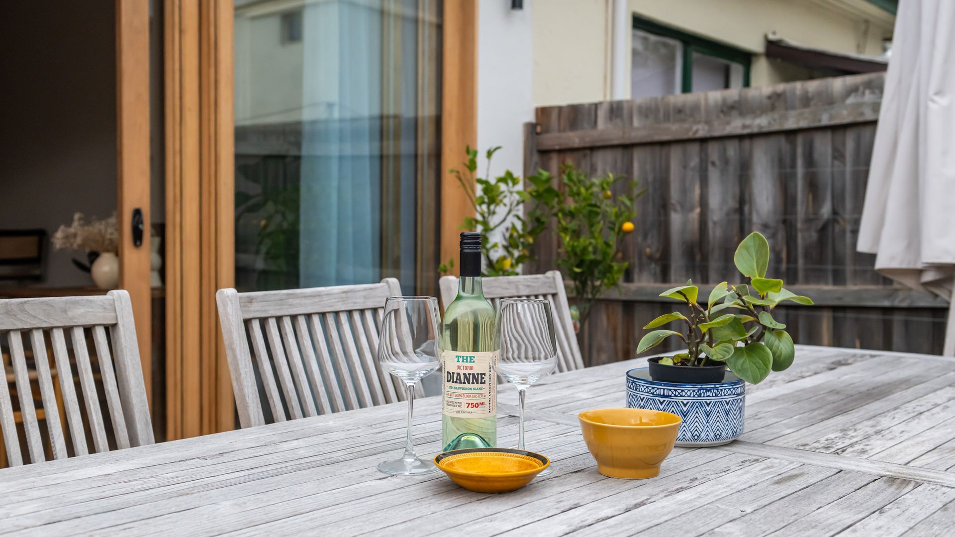 Una mesa de madera con una botella de vino, dos copas, un bol amarillo y una planta en maceta.