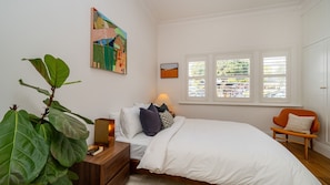 A bedroom with a bed, bedside table, lamp, chair, large plant, and artwork on the wall. - Bellamy Richmond by Property Mums (Richmond)