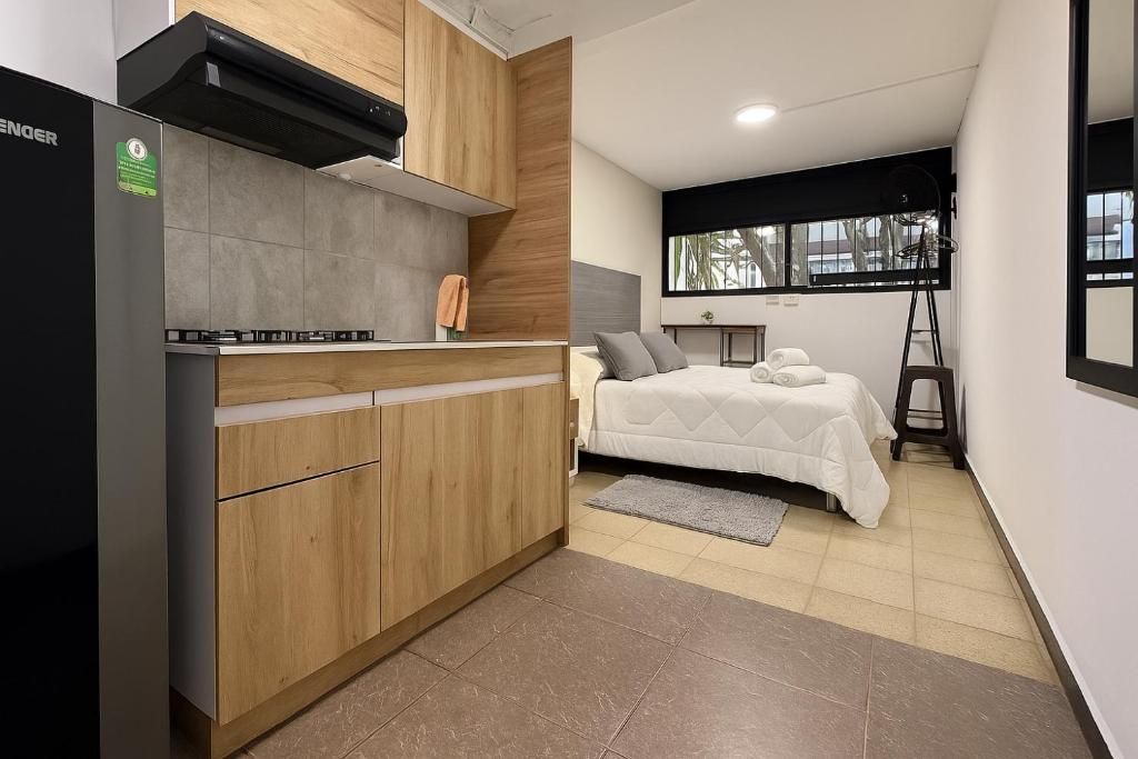 Cocina compacta con estufa integrada y armarios de madera, cama con cubrecama blanco y ventana que deja entrar luz natural.