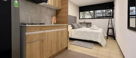 Cocina compacta con estufa integrada y armarios de madera, cama con cubrecama blanco y ventana que deja entrar luz natural.