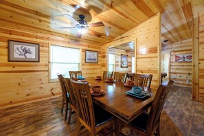 Une salle à manger en bois, avec une grande table et des chaises, des tableaux encadrés aux murs et un ventilateur de plafond.