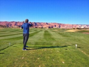 A person standing on a golf course with a clear sky and mountains in the background.