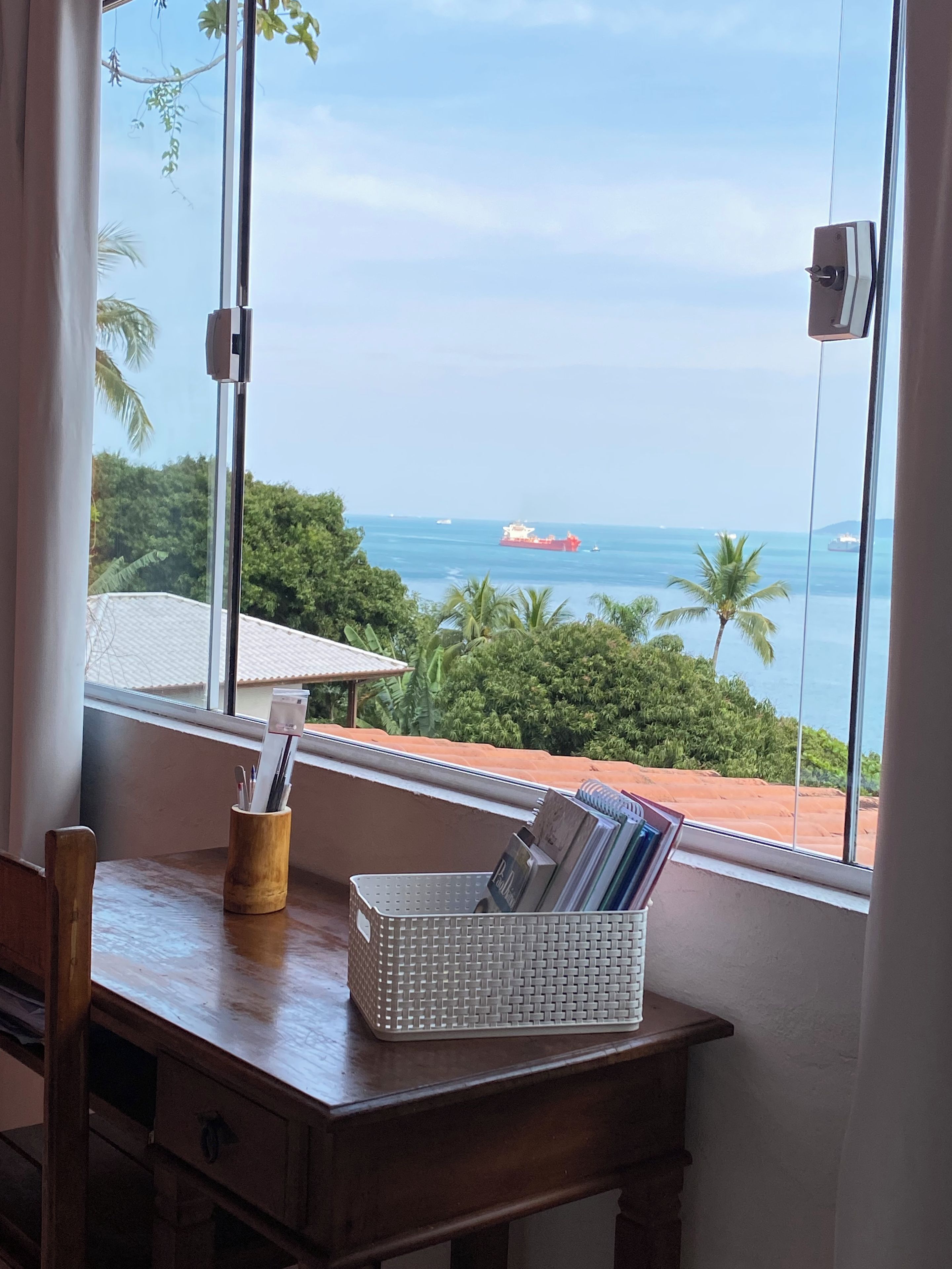 Un bureau en bois, sur lequel se trouve un panier blanc rempli de magazines, est placé près d’une fenêtre donnant sur une vue côtière, avec des palmiers et un navire rouge visible au loin.