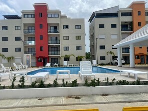 A swimming pool with lounge chairs and a building with multiple balconies. - APARTAMENTO COMPLETO A 2 MINUTOS DE LA PLAYA CON PISCINA 3 DORMITORIOS 2 BAÑOS (La Romana)