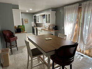 A dining area with a wooden table and chairs, a kitchen in the background, and a window with sheer curtains. - Gorgeous 4-bedroom house with AC in charming Potomac (Potomac)