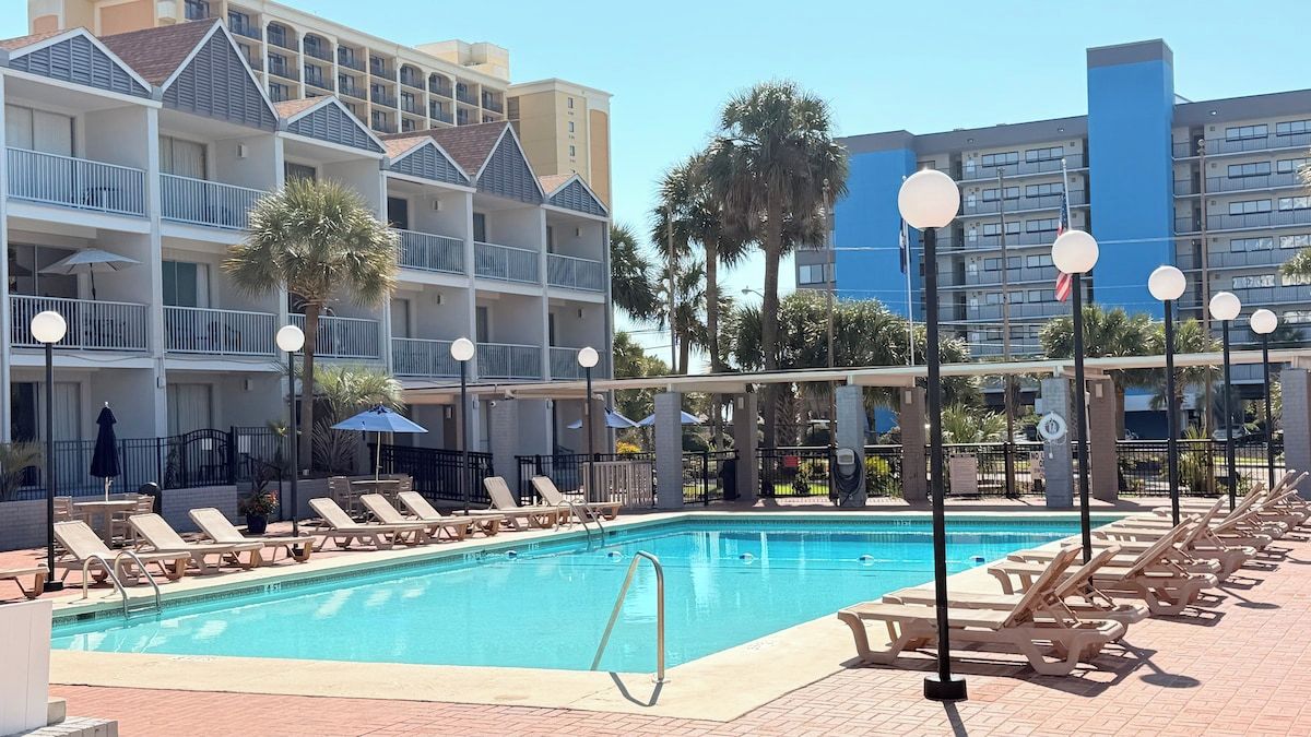 Ein Swimmingpool mit Liegestühlen, einer Palme und einem Hotel im Hintergrund.