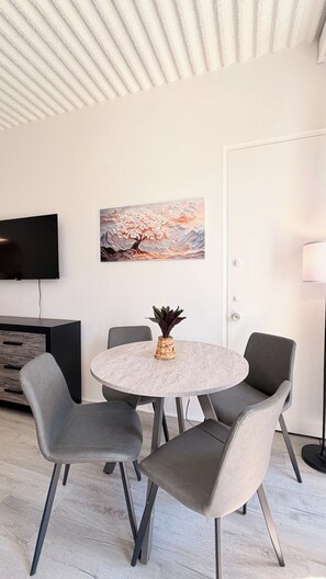 A modern living room with a round table, grey chairs, a flat-screen TV, a painting, and a lamp. - Cozy Beach Studio | Sleeps 4 | First floor (Myrtle Beach)
