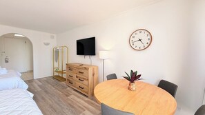 Une chambre d’hôtel avec un lit, une commode en bois, un téléviseur, une table ronde avec des chaises et une horloge murale.
