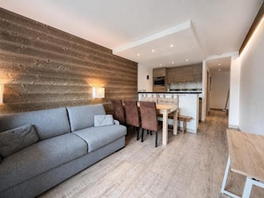 A modern living room with a grey sofa, wooden wall paneling, and a dining area with a wooden table and chairs. - 20m from ski slopes, 1st floor, balcony, tv, ski locker, 35m², Les Menuires (Les Menuires)