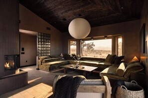 A modern living room with a fireplace, a large window with a view, and a cozy seating area. - Solace House · Borders JT Park, Pool, Hot Tub (Joshua Tree)