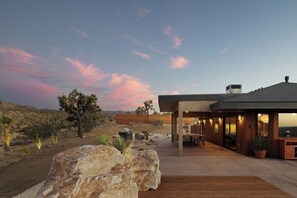 A modern outdoor patio with a dining area, large rock feature, and a view of the desert landscape. - Solace House · Borders JT Park, Pool, Hot Tub (Joshua Tree)