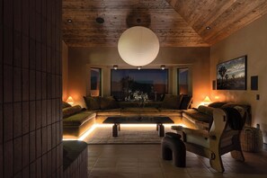 A cozy living room with a large sofa, a coffee table, and a hanging light fixture. - Solace House · Borders JT Park, Pool, Hot Tub (Joshua Tree)