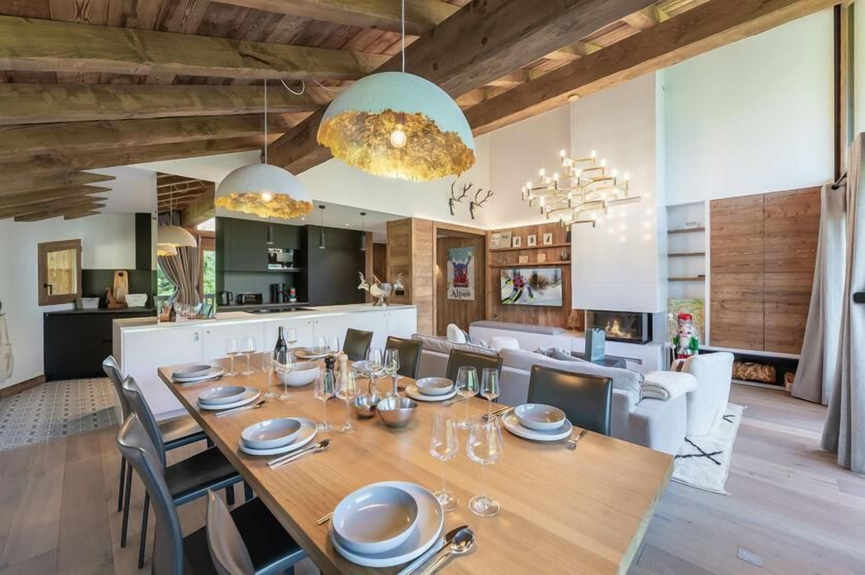 Un comedor amplio con una larga mesa de madera dispuesta para una comida, una cocina al fondo y una moderna lámpara de araña.