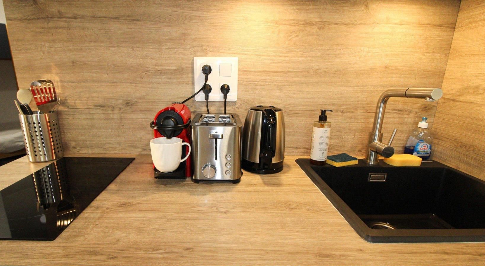 A kitchen with a coffee maker, toaster, kettle, and a sink with cleaning supplies.