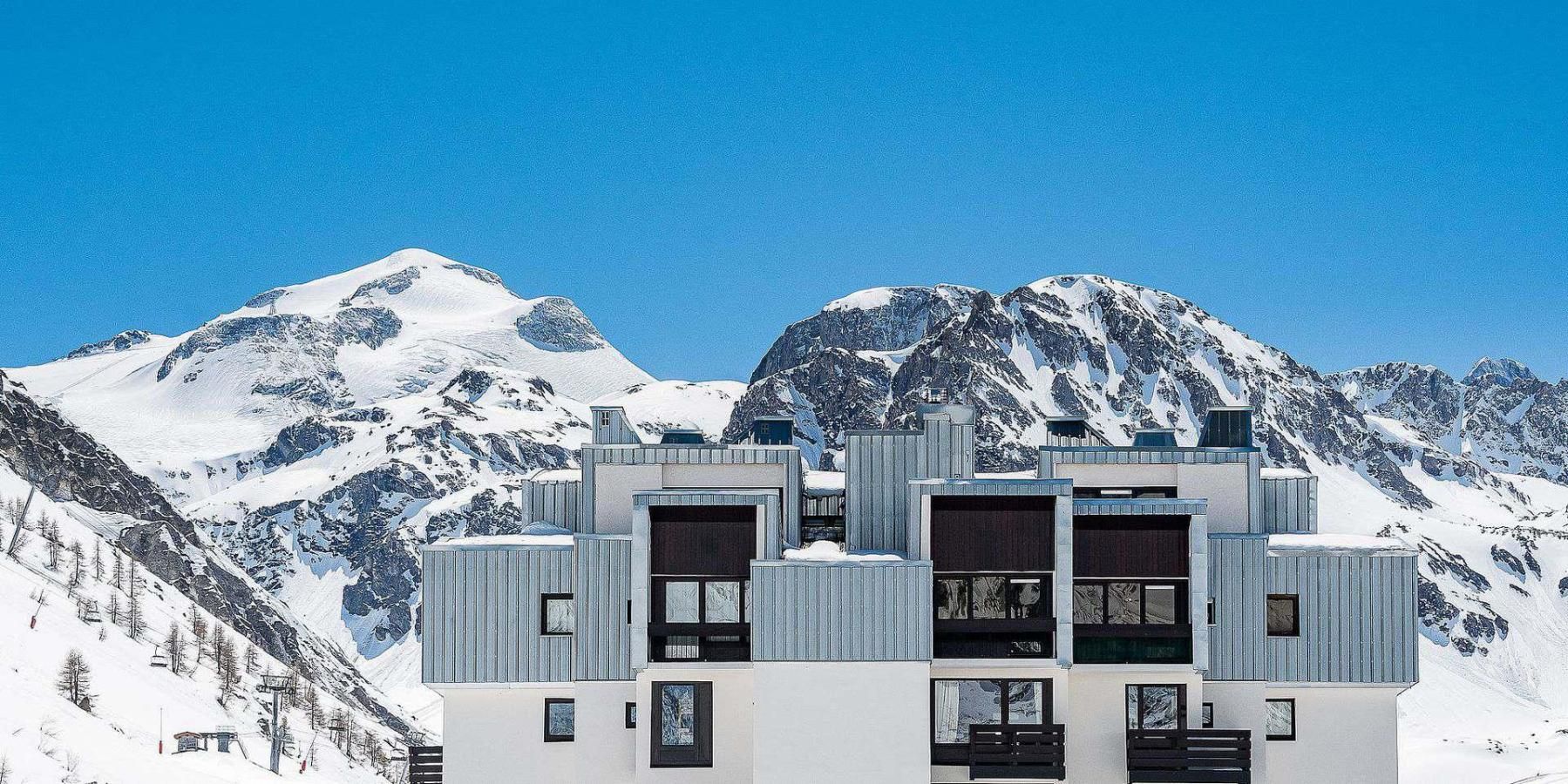 A modern building with a symmetrical facade, large windows, and a flat roof, set against a backdrop of snow-covered mountains.