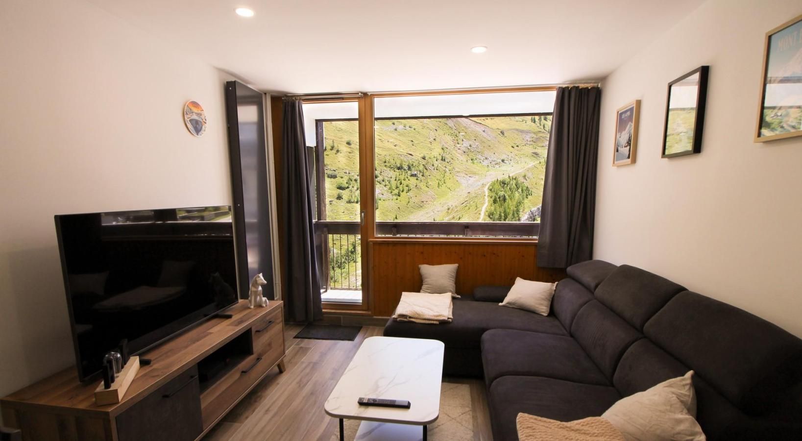 Ett modernt vardagsrum med en platt-TV, en soffa i mörk färg, ett litet bord och ett stort fönster med utsikt över ett grönskande landskap.