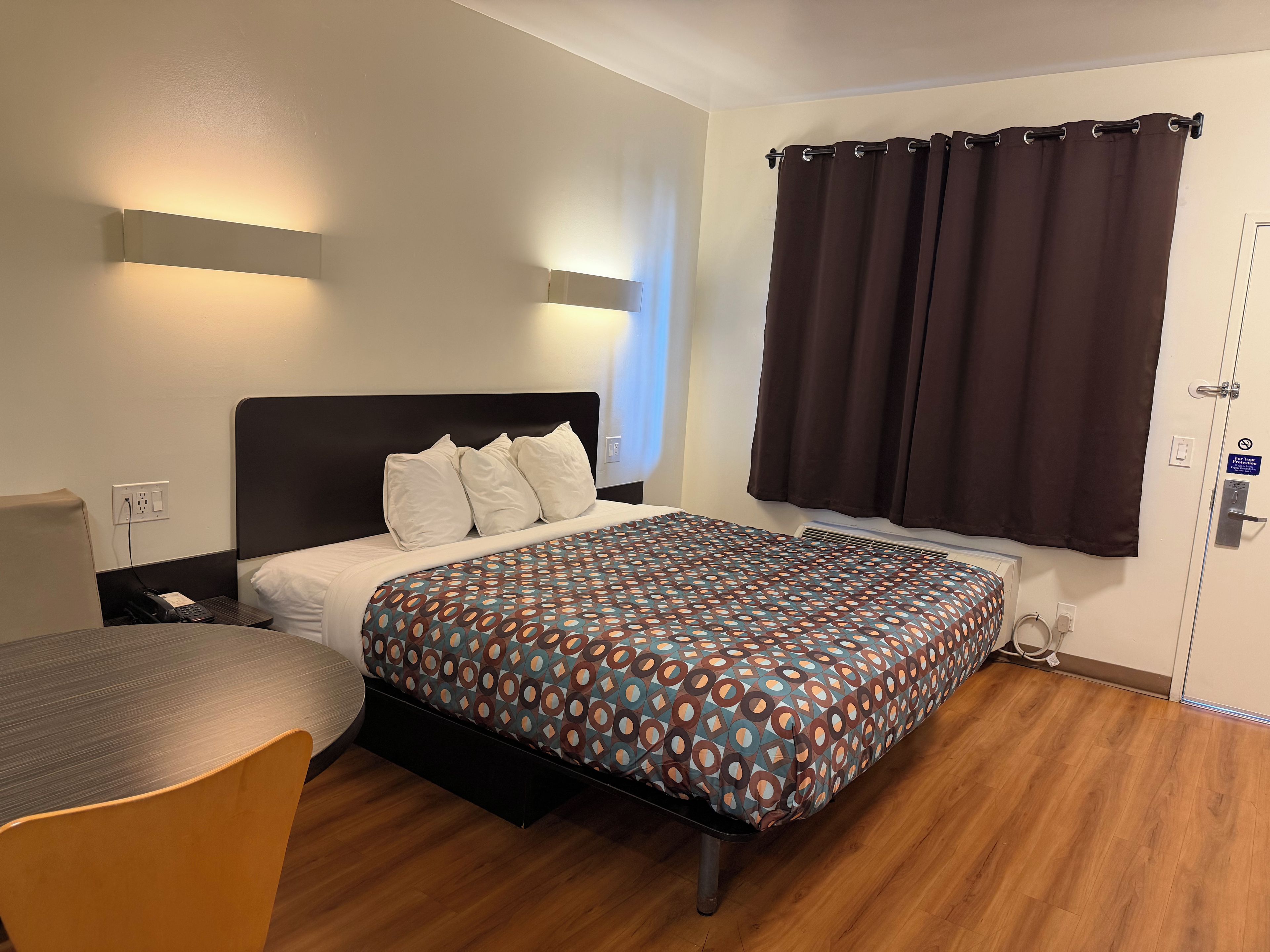 A hotel room with a bed, bedside table, chair, and a window with curtains.