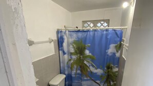 Kylpyhuone, jossa on suihkuverho, jossa on kuvattuna palmuja ja pilviä, wc-istuin, pesuallas ja sininen roskakori.