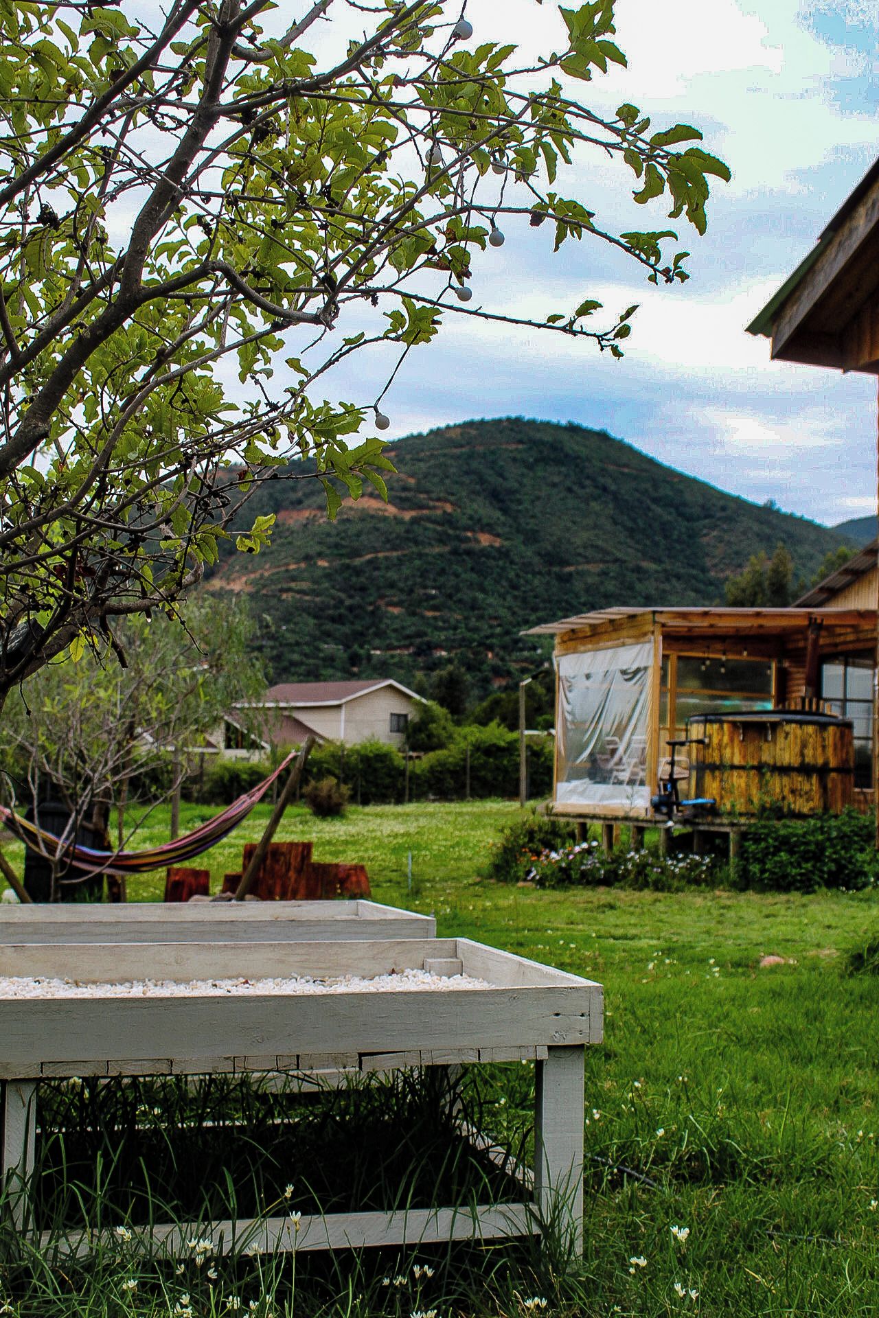 A wooden deck with a hammock, a wooden table, and a view of a mountain and a house in the background.