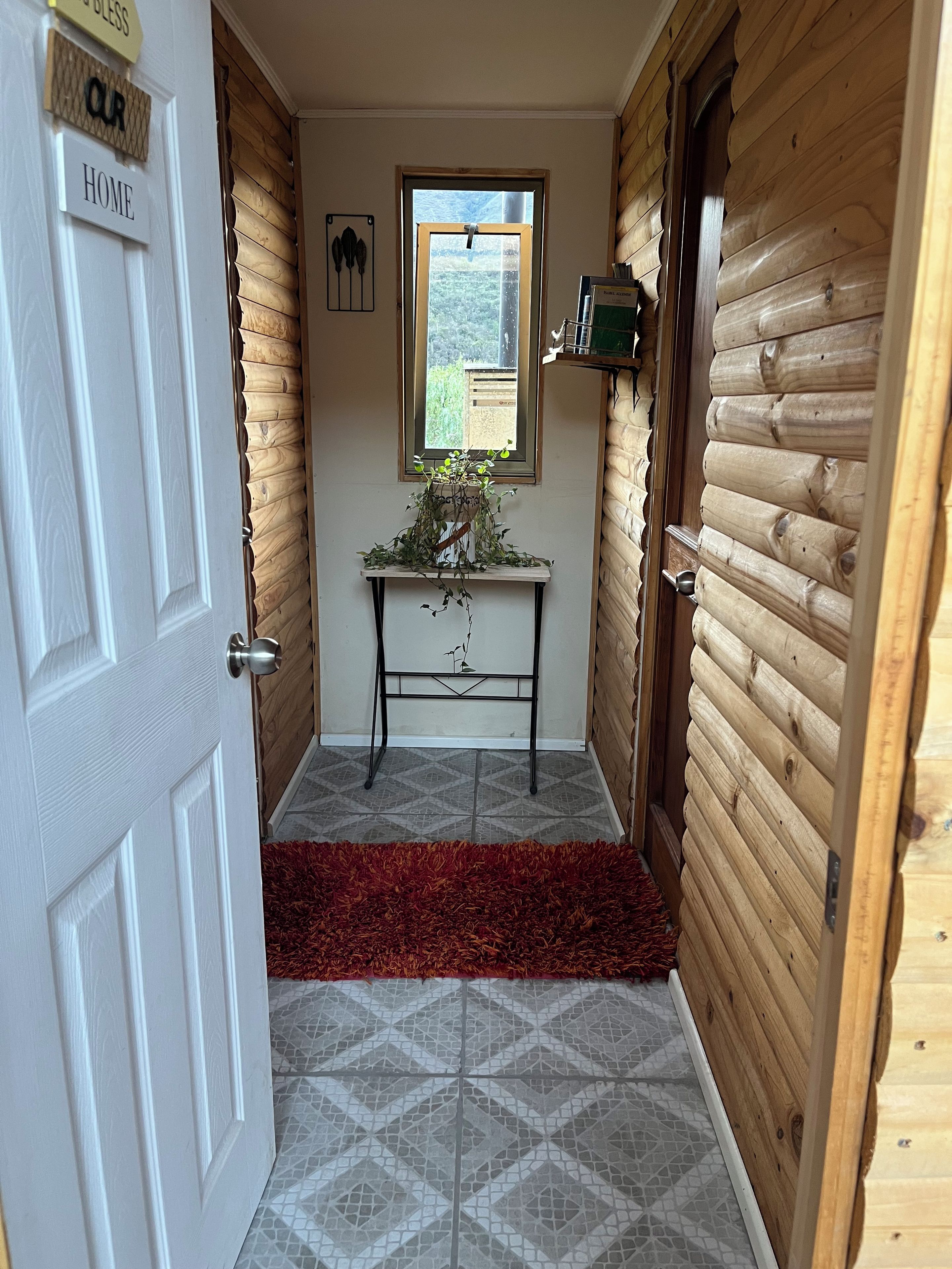 A hallway with a wooden door labeled 'OUR HOME,' a small table with a plant, and a window with a view.