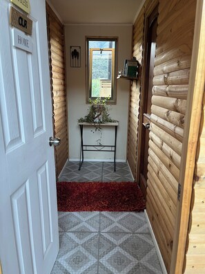 Un couloir avec une porte en bois portant l’inscription « NOTRE FOYER », une petite table avec une plante et une fenêtre donnant sur un paysage.