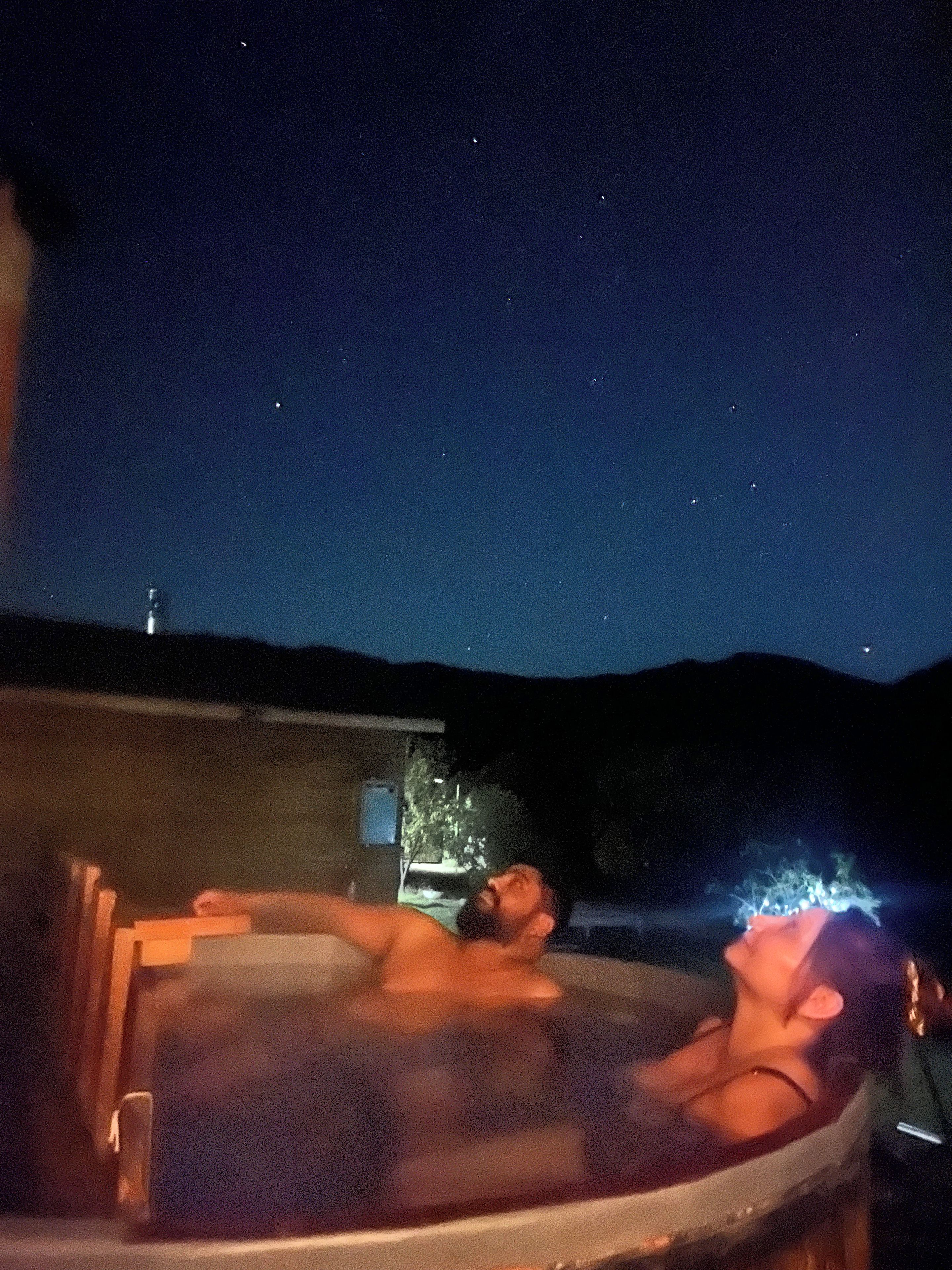 A person relaxing in a hot tub outdoors at night with a clear view of the sky and distant mountains.