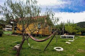 Uma cabana de madeira com uma rede em uma área gramada, rodeada por árvores e uma montanha ao fundo.