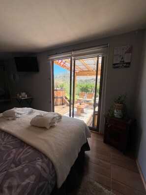 A bedroom with a bed, a TV, a sliding glass door to an outdoor patio with a wooden deck and chairs.