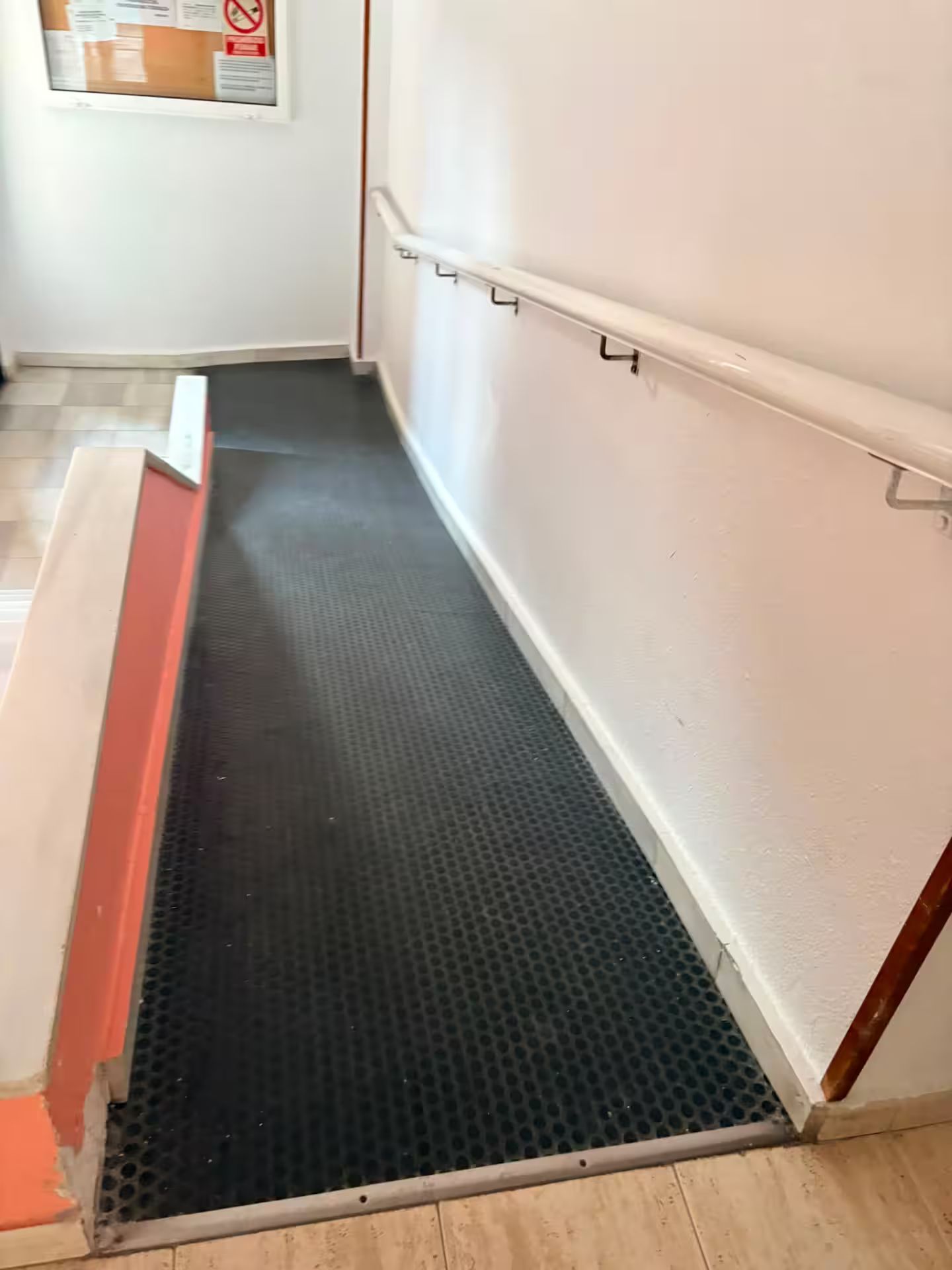 A narrow, enclosed space with a textured floor, a white wall, and a wooden floor visible at the bottom.