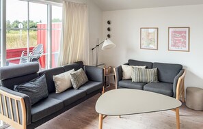 Een woonkamer met een grijze bank, een ronde salontafel en een rood terras zichtbaar door het raam.