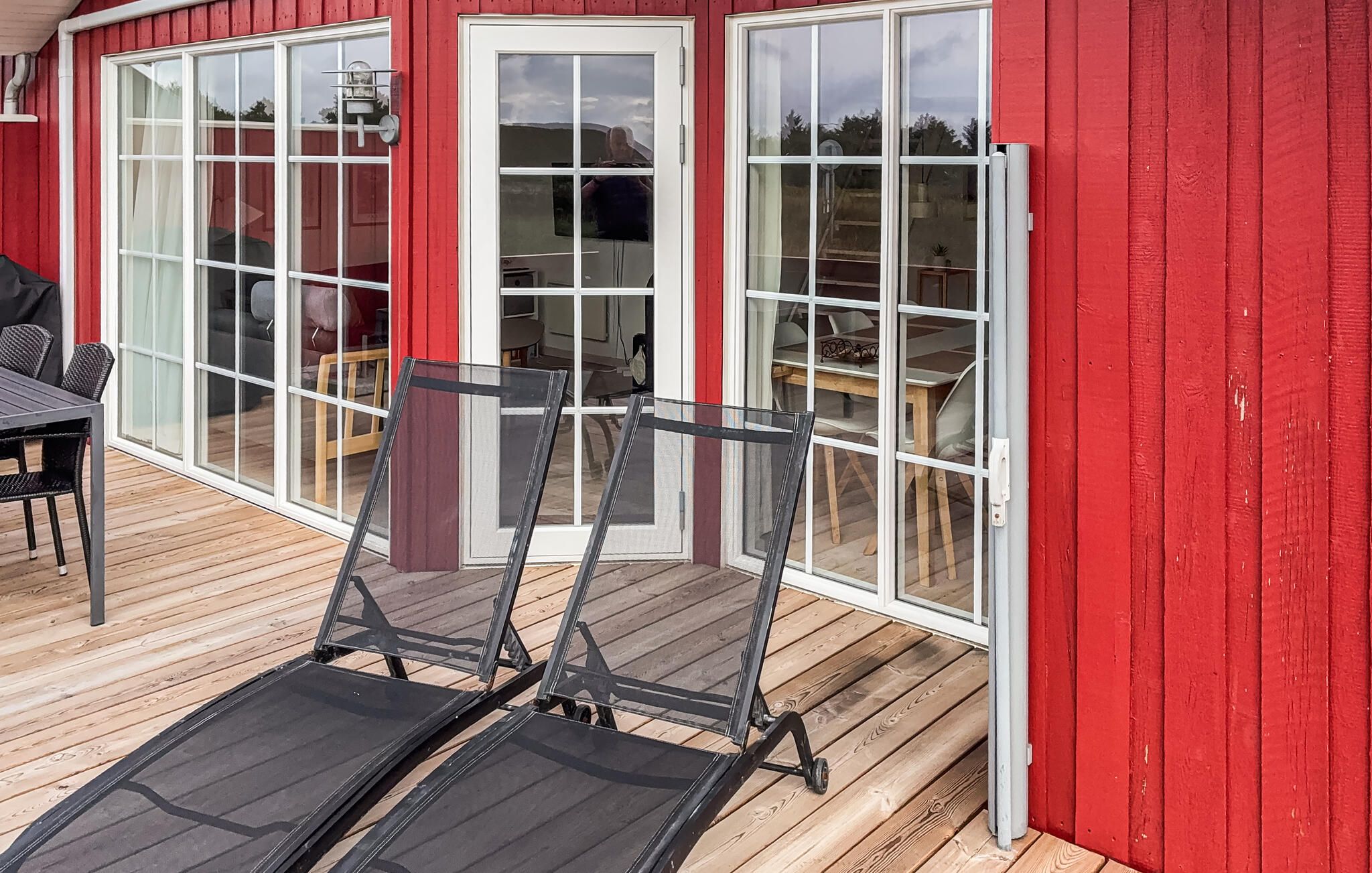 Un edificio rojo con amplios ventanales y puertas de vidrio, una terraza de madera y una silla de metal.