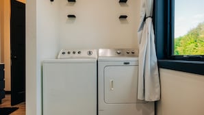 Une buanderie équipée d’un lave-linge et d’un sèche-linge, une fenêtre donnant sur un espace verdoyant et une porte menant à une autre pièce.