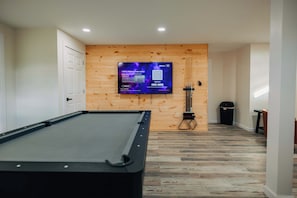 A pool table in a room with wooden panel walls, a mounted TV, and a trash bin.