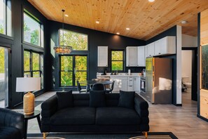 A modern interior with a black sofa, dining table, and chairs, a kitchen with white cabinets, and large windows offering a view of greenery. - The Overlook A Luxury Cabin With Hot Tub And Views (New Plymouth)