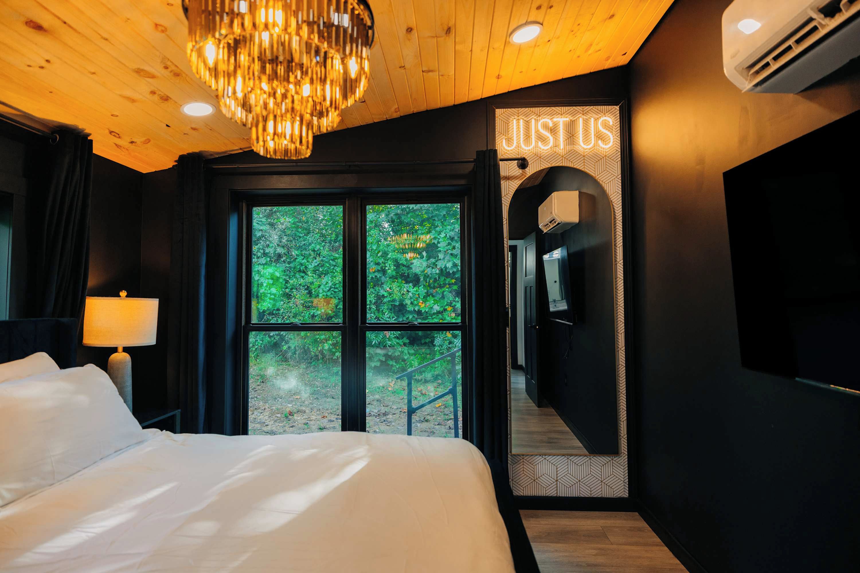 Habitación de hotel con cama, candelabro, ventana con vistas a zonas verdes y pasillo con un cartel que dice 'JUSTUS'.
