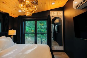 Une chambre d’hôtel avec un lit, un lustre, une fenêtre donnant sur un paysage verdoyant et un couloir sur lequel est affiché un panneau indiquant « JUSTUS ».