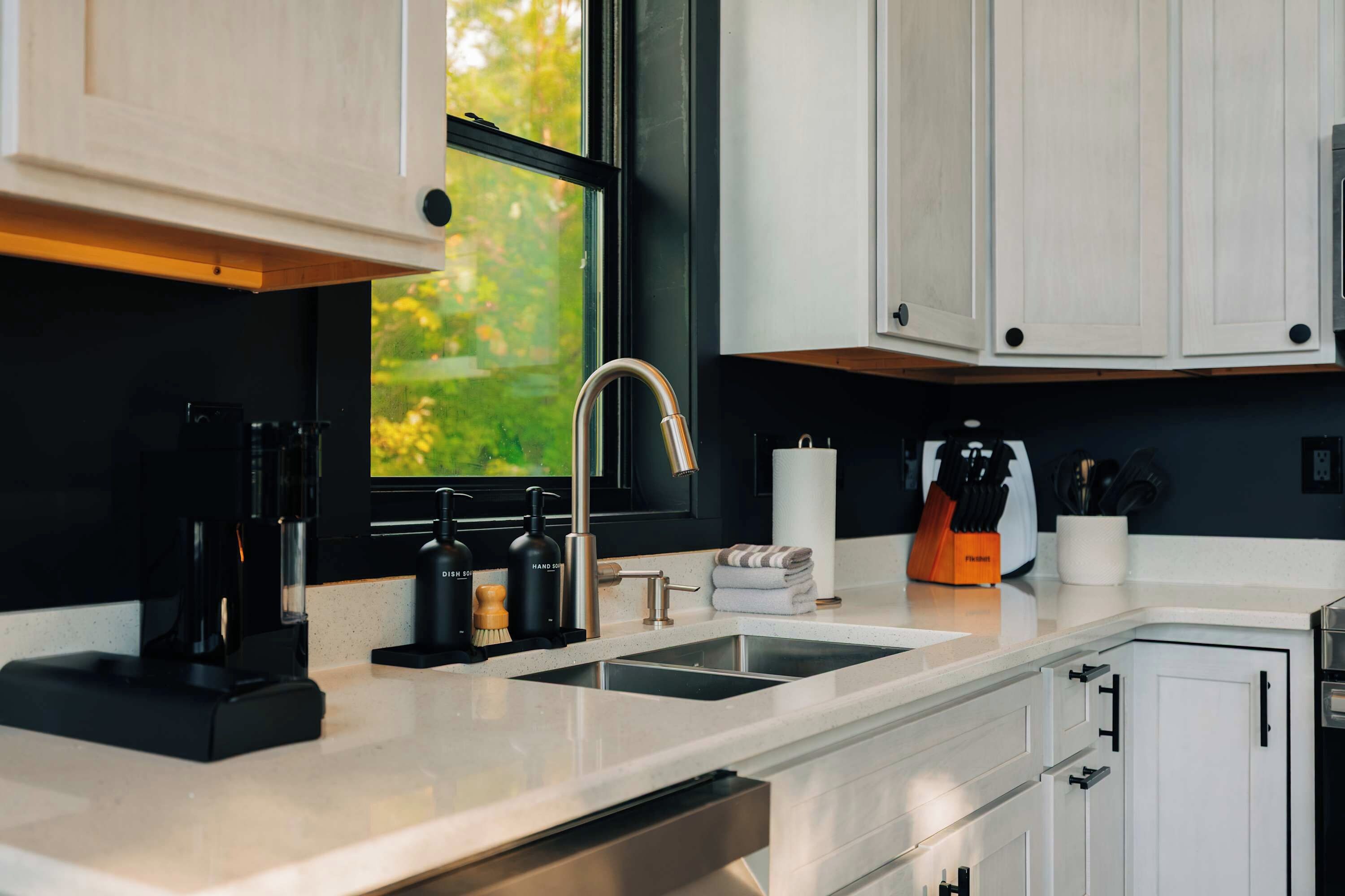 A modern kitchen with a stainless steel sink, a coffee machine, and a window with a view of greenery.