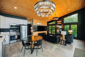 A modern kitchen with stainless steel appliances, a dining area with a round table and chairs, a black sofa, and a large window with a view of greenery. - The Overlook A Luxury Cabin With Hot Tub And Views (New Plymouth)