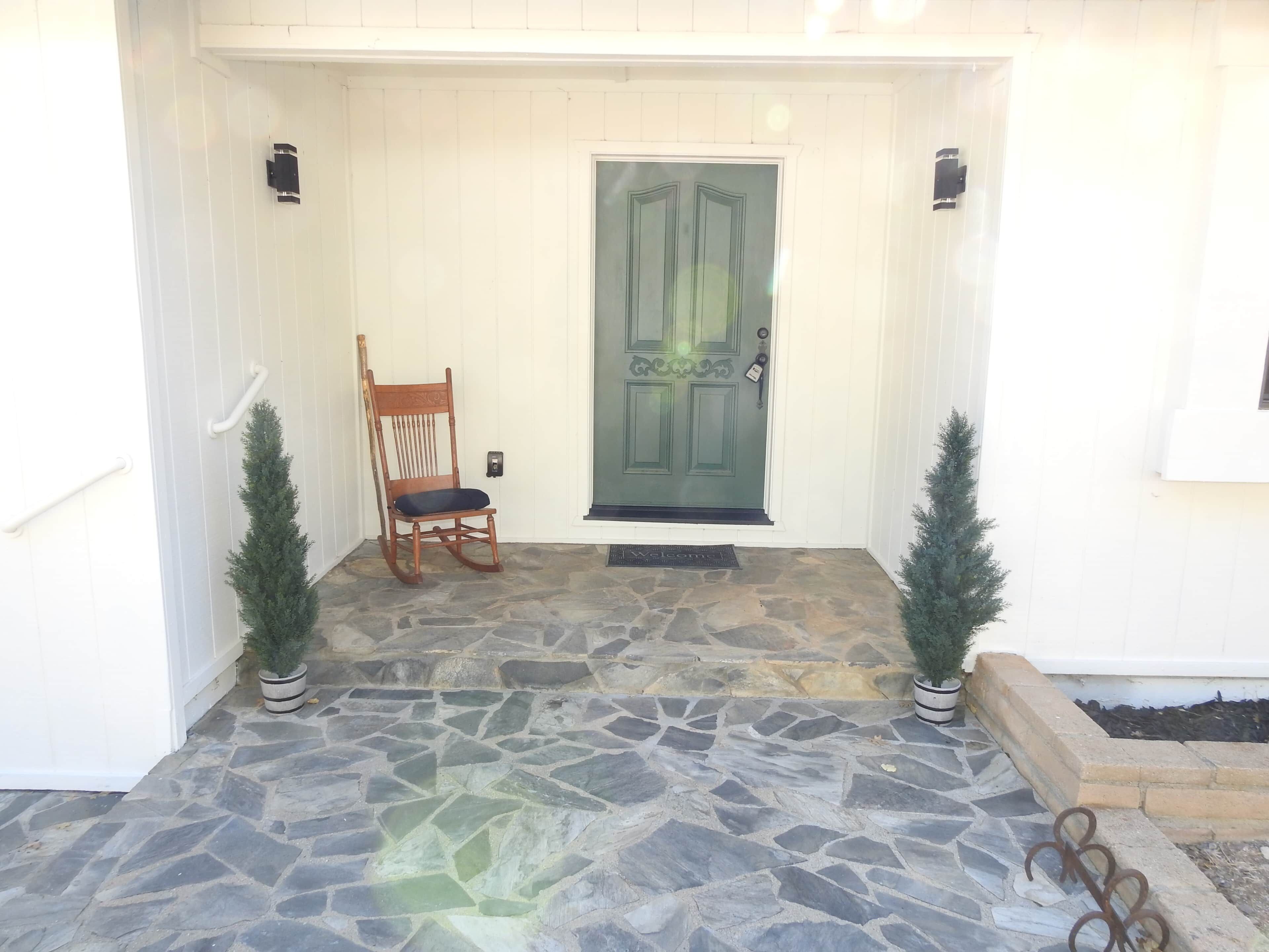 Entrada com calçada de pedra, uma cadeira de balanço de madeira e dois vasos de plantas.