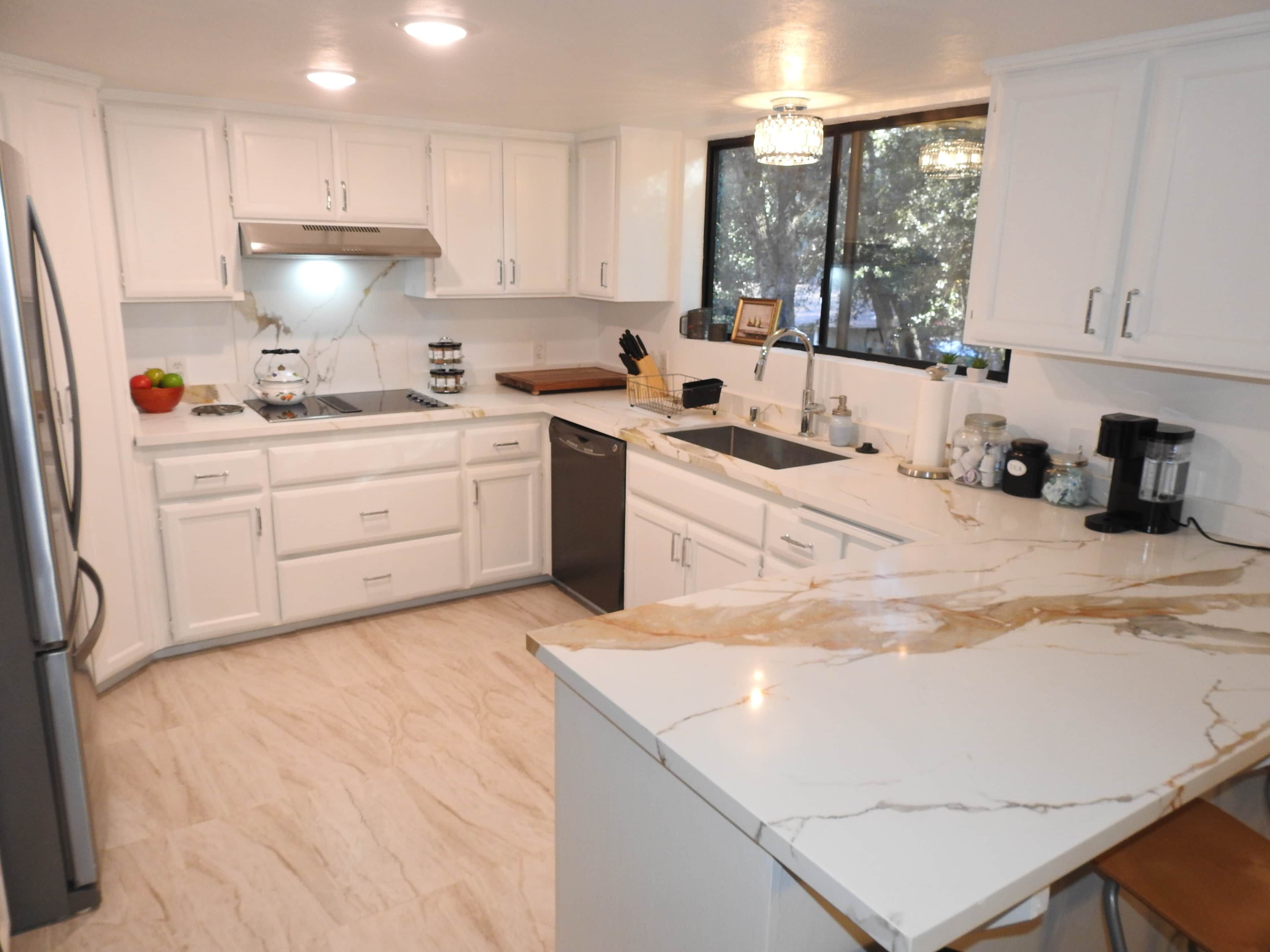 A modern kitchen with white cabinetry, a marble countertop, and a window providing a view of trees.