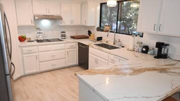 A modern kitchen with white cabinetry, a marble countertop, and a window providing a view of trees.