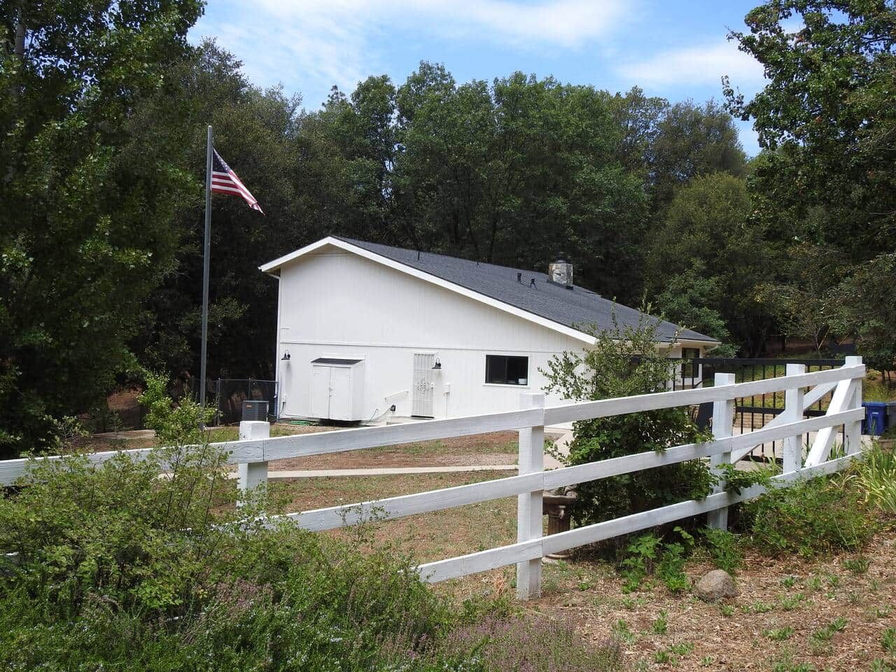 A white barn with a flagpole, fenced area, and trees in the background.