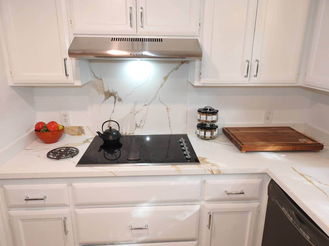 A kitchen with white cabinets, a stainless steel range hood, a black induction cooktop, a wooden cutting board, and a bowl of fruit.