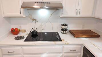 A kitchen with white cabinets, a stainless steel range hood, a black induction cooktop, a wooden cutting board, and a bowl of fruit.