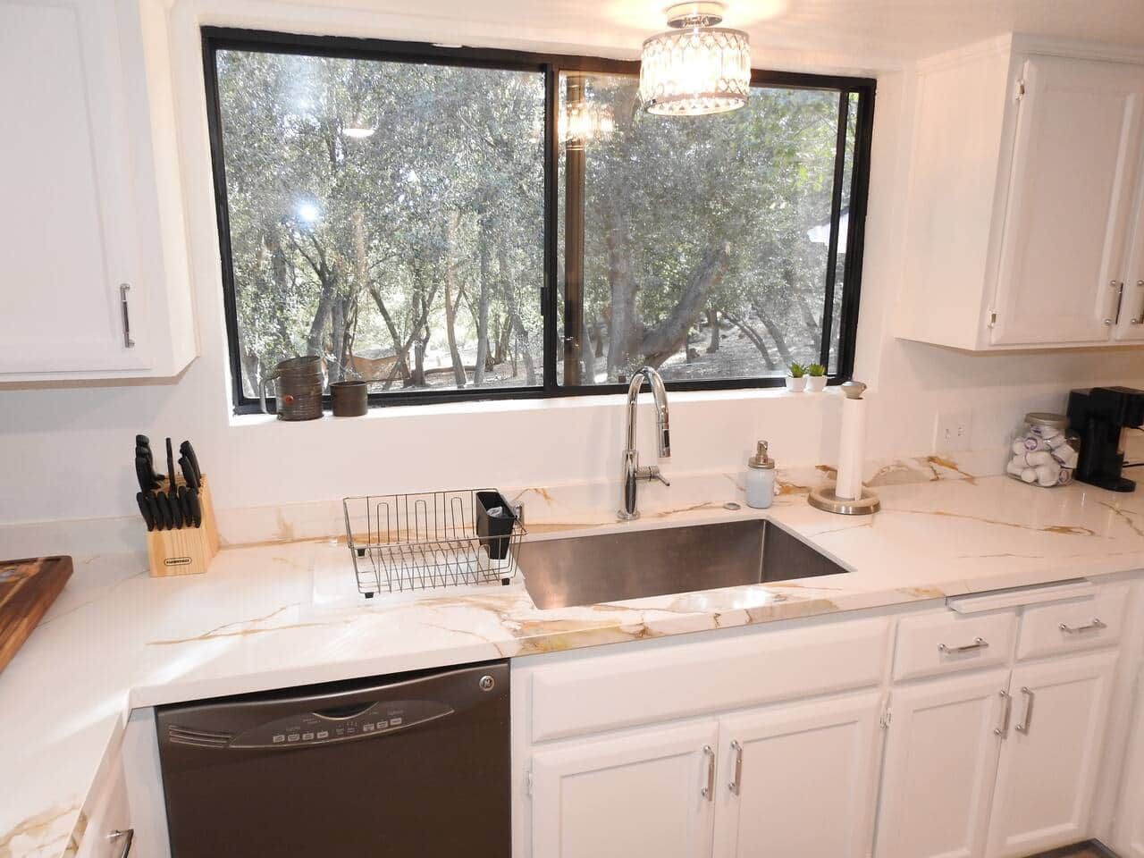Una cocina con un lavamanos doble, armarios blancos, lavavajillas y una ventana con vistas a árboles.